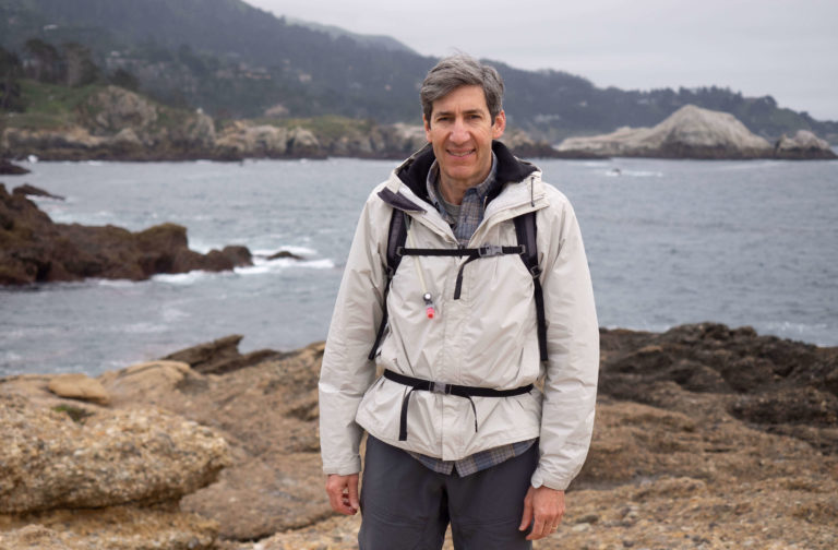 Dave Heiner standing on a rocky coastline wearing a light hiking jacket and backpack, with ocean waves and cliffs in the background.