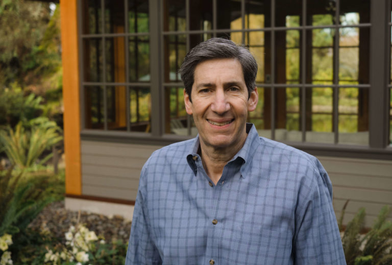 Dave Heiner smiling outdoors in front of a building with large windows, wearing a blue checkered shirt.