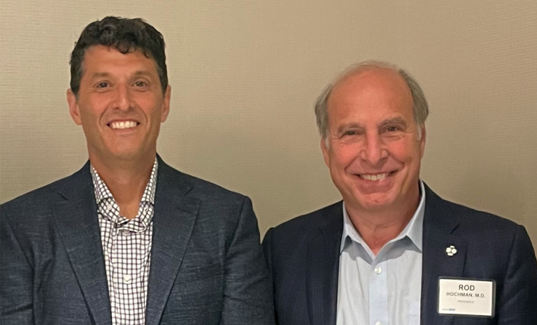 Two men standing side by side, both wearing jackets over collared shirts, smiling in front of a neutral-colored wall.