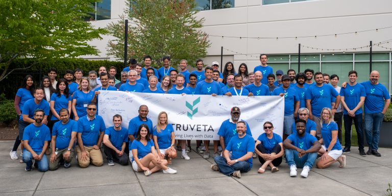 Large group of people wearing blue Truveta shirts posing outdoors in front of an office building, holding a Truveta banner signed with messages.