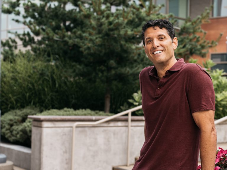 Terry Myerson, Truveta’s CEO, smiling outdoors in a maroon shirt, leaning on a concrete railing with greenery and modern buildings in the background.