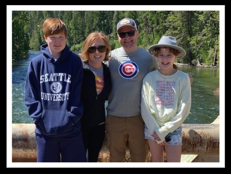 A family of four stands together on a sunny day by a forested river, smiling at the camera, wearing casual clothes including a hoodie, hat, sunglasses, and T-shirt.