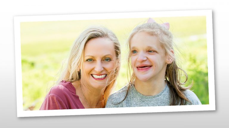 Two smiling women outdoors, one with blonde hair and one with a pink bow, enjoying a bright sunny day together in a grassy field.