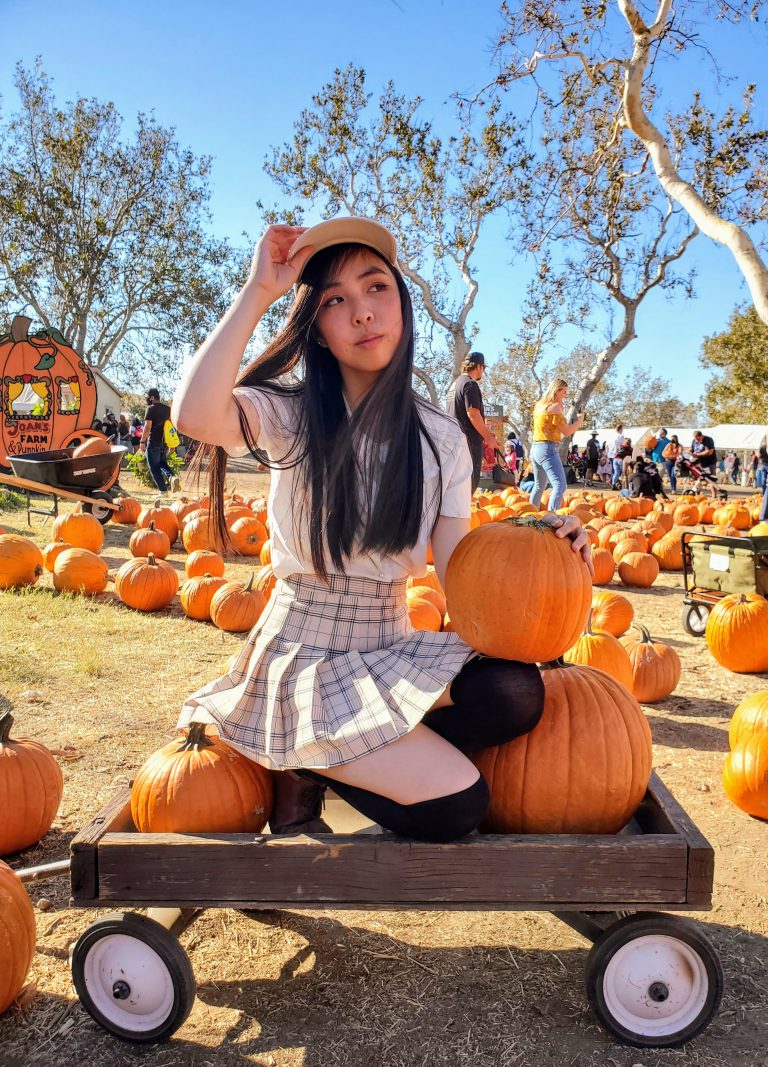 Person sitting on a wooden wagon at a pumpkin patch, holding a pumpkin and adjusting their hat, surrounded by bright orange pumpkins under a clear blue sky.