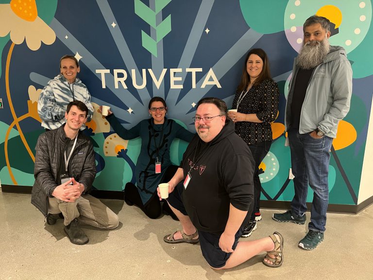 Group of Truveta team members posing and smiling in front of a colorful mural with the Truveta logo, holding small cups during an office gathering.
