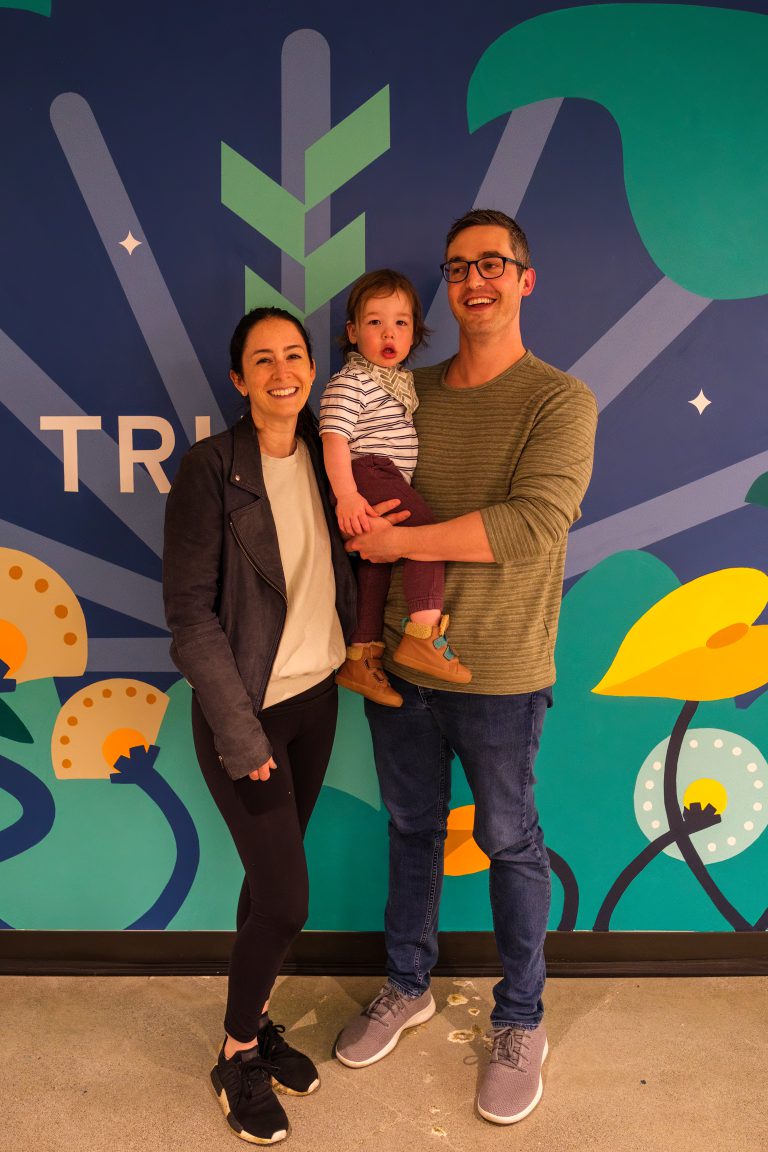 A smiling family stands together in front of a colorful Truveta mural, with one adult holding a toddler as they pose cheerfully in the bright, playful space.