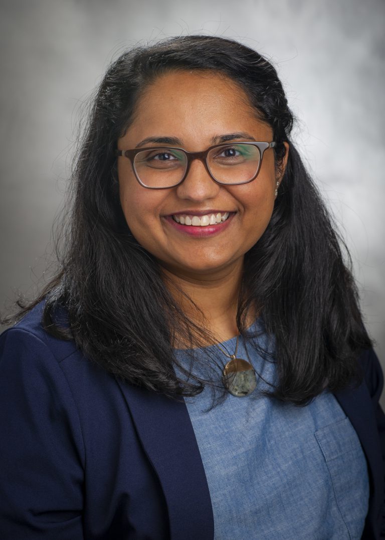 Professional headshot of a woman, smiling at the camera while wearing glasses, a blue top, and a navy blazer, with a soft gray background behind her.