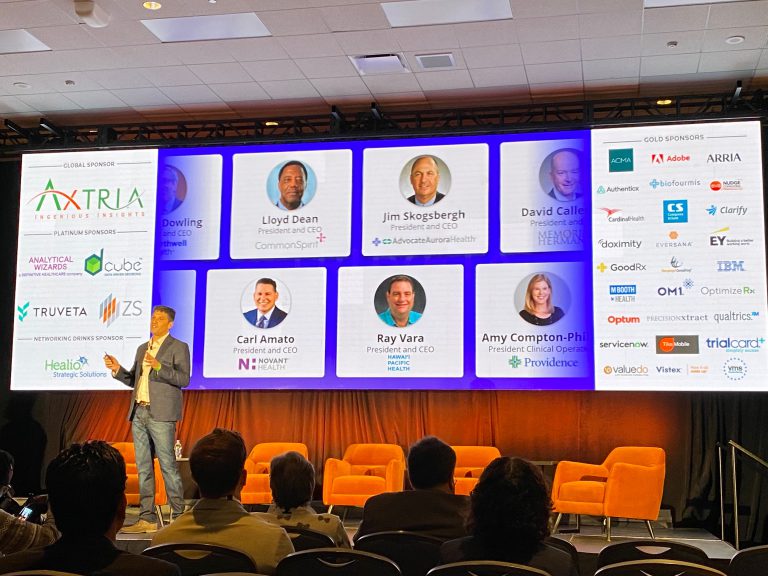 Terry Myerson, CEO of Truveta, presents on stage at a healthcare leadership event, with sponsor logos and executive headshots displayed on a large screen behind him.