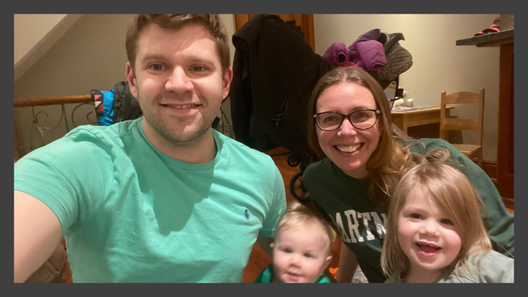 Smiling family of four taking a selfie indoors, including two adults and two young children.