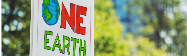 Hand-drawn sign reading ‘One Earth’ with an illustrated globe, photographed outdoors against a blurred green background.