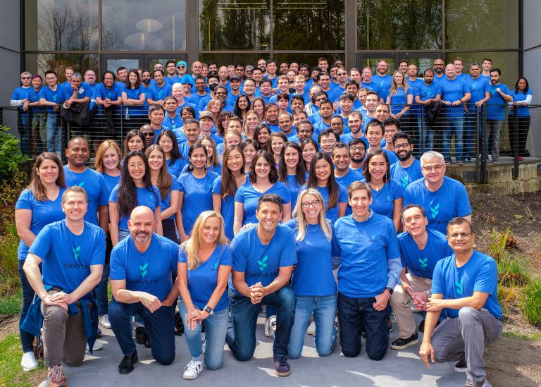 Large group of people wearing matching blue shirts pose together outdoors in front of a building for a team photo.
