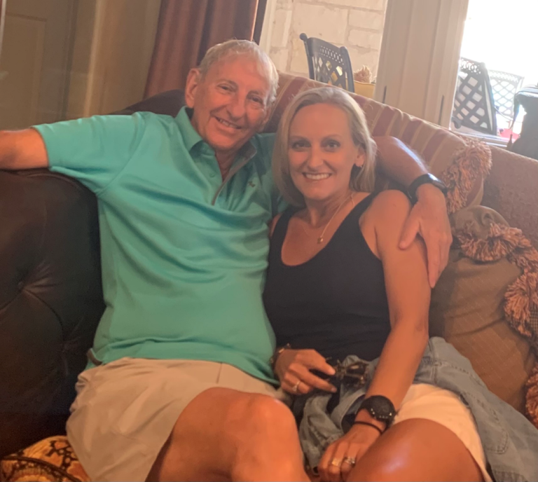 Two people sitting closely on a couch indoors, smiling and posing together in a relaxed home setting.