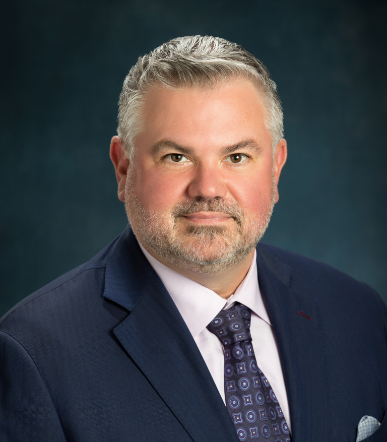 Professional headshot of a man wearing a suit and tie, facing the camera against a dark studio background.