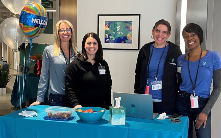 Four Truveta employees stand side by side behind a welcome table in an office lobby, smiling at the camera, with balloons, snacks, and a laptop visible.