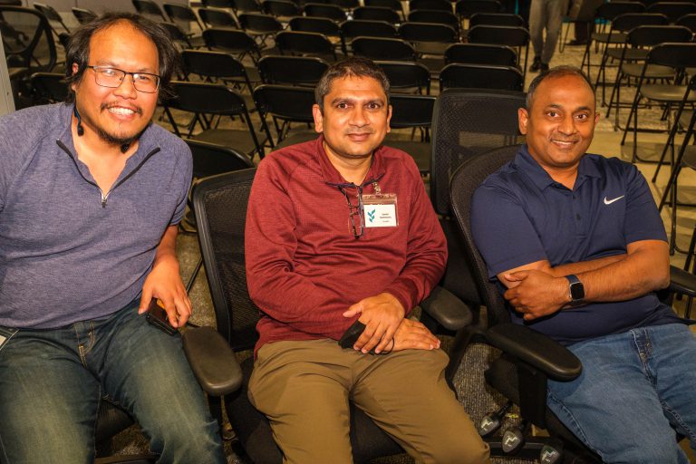Three colleagues seated in conference chairs, smiling toward the camera in a large meeting room with rows of empty chairs behind them.