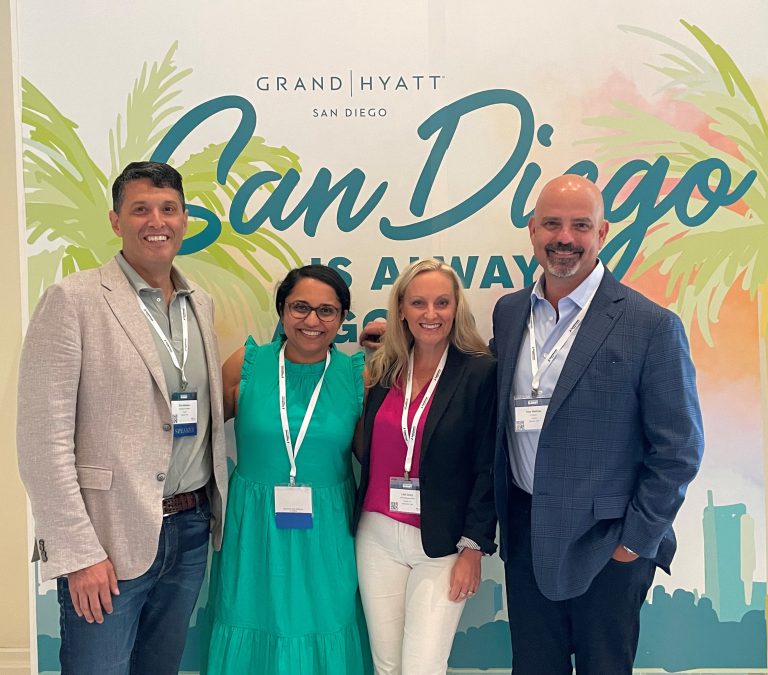 Four professionals pose at a Grand Hyatt San Diego conference, smiling in front of a Sam Diego themed event backdrop.