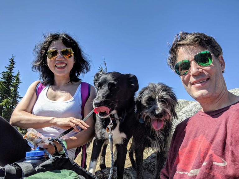 Two adults and two dogs pause on a sunny mountain hike, smiling at the camera against a clear blue sky.