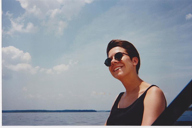 A picture of a woman in a sunglasses and a black top, smiling, looking at the ocean.