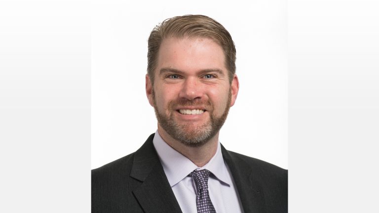 Professional headshot of a smiling man with short light-brown hair and a trimmed beard, wearing a dark suit, light dress shirt, and patterned tie on a white background.