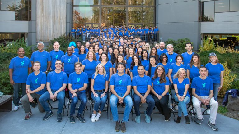 Large group of Truveta employees wearing blue Truveta shirts pose together outside an office building, representing the company team and culture.