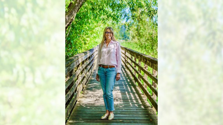 Woman wearing glasses, a light pink shirt, and jeans stands smiling on a wooden footbridge surrounded by lush green trees and sunlight.