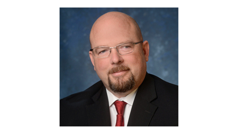 Professional headshot of a bald man wearing glasses, a dark suit, white shirt, and red tie, facing the camera with a calm expression against a blue studio background.