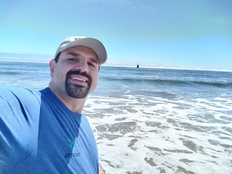 Smiling person wearing a Truveta T-shirt and baseball cap takes a selfie at the beach, with ocean waves, blue sky, and a sailboat in the distance.