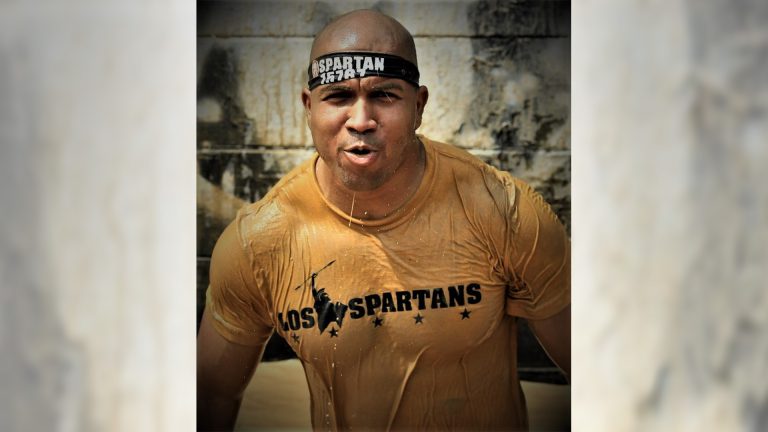 Athletic man wearing a Spartan headband and wet tan T-shirt stands mid-exertion, covered in water and mud during an outdoor obstacle race.