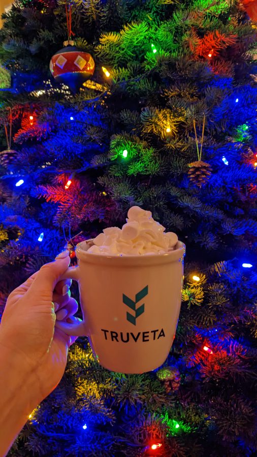 A hand holding a Truveta mug of cocoa in front of a decorated Christmas tree.