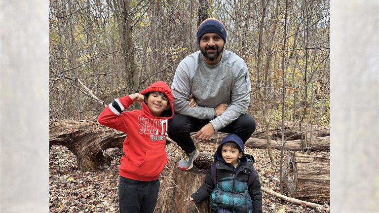 Adult and two children pose on a tree stump in a wooded area during fall, with one child flexing an arm and leaves covering the forest floor.